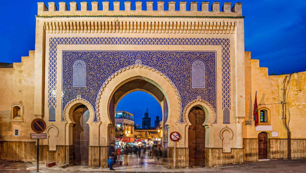 Grand crenellated entrance gate to a Moroccan city is covered in blue geometric tiles in front of bright blue sky.