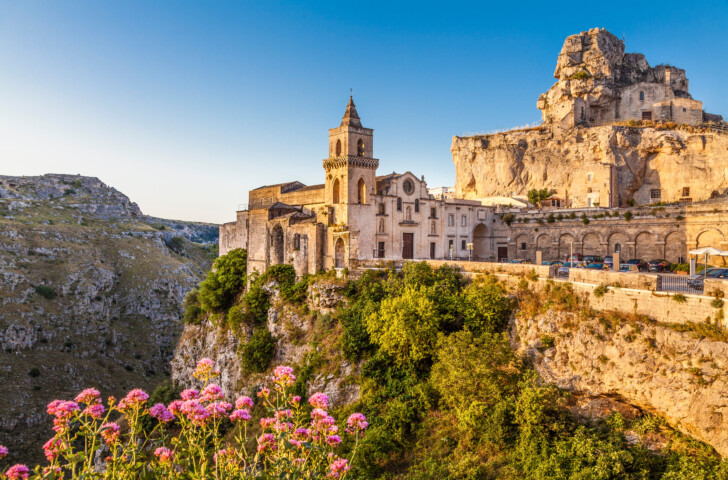 A historic stone-built church and buildings sit on a cliffside in Matera, Italy, with a backdrop of rugged terrain and vibrant pink flowers in the foreground on a sunny day.