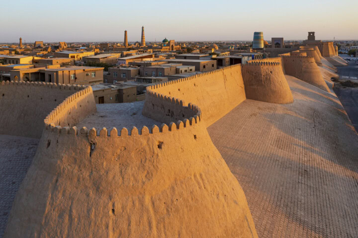 A panoramic view of ancient city walls and towers at sunset, with buildings and minarets visible in the background, evokes the rich history of Central Asia. The mesmerizing scene captures the essence of Uzbekistan, making it a traveler's dream destination.