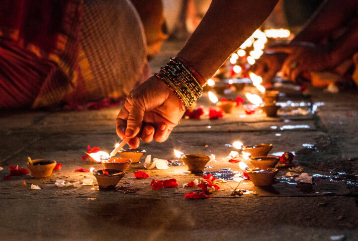 A hand holding a match lights an earthen lamp among a row of glowing lamps and scattered flower petals during a cultural or religious ceremony in Nepal.