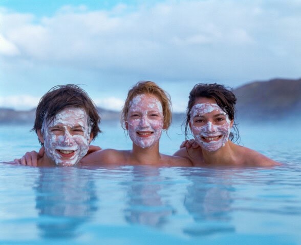 Three people with white face masks smile while partially submerged in a body of water, with a cloudy sky and Iceland's mountainous landscape in the background, capturing the essence of travel and tourism to this stunning destination.