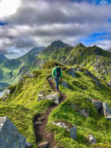 A person in a green jacket hikes along a narrow, rocky trail on a green mountain ridge under a partly cloudy sky, capturing the rugged beauty of Norway.