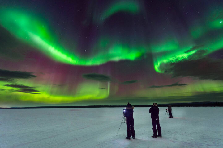 Three people stand on a snowy landscape in Finland at night, observing and photographing a vibrant green and purple aurora borealis display in the sky, capturing the magic of Arctic tourism.