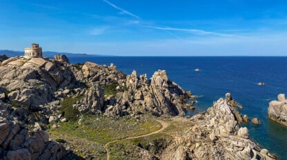 A rocky coastal landscape with a small circular building on a hill overlooking the sea under a clear blue sky, reminiscent of the adventures one might find in Corsica or Sardinia.