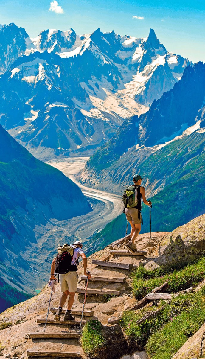 Two hikers with backpacks and trekking poles ascend stone steps on a mountain trail, with a rugged mountain range and a glacier visible in the background under a clear blue sky. Incorporate these stunning scenes into your travel blog for improved SEO by using relevant keywords.