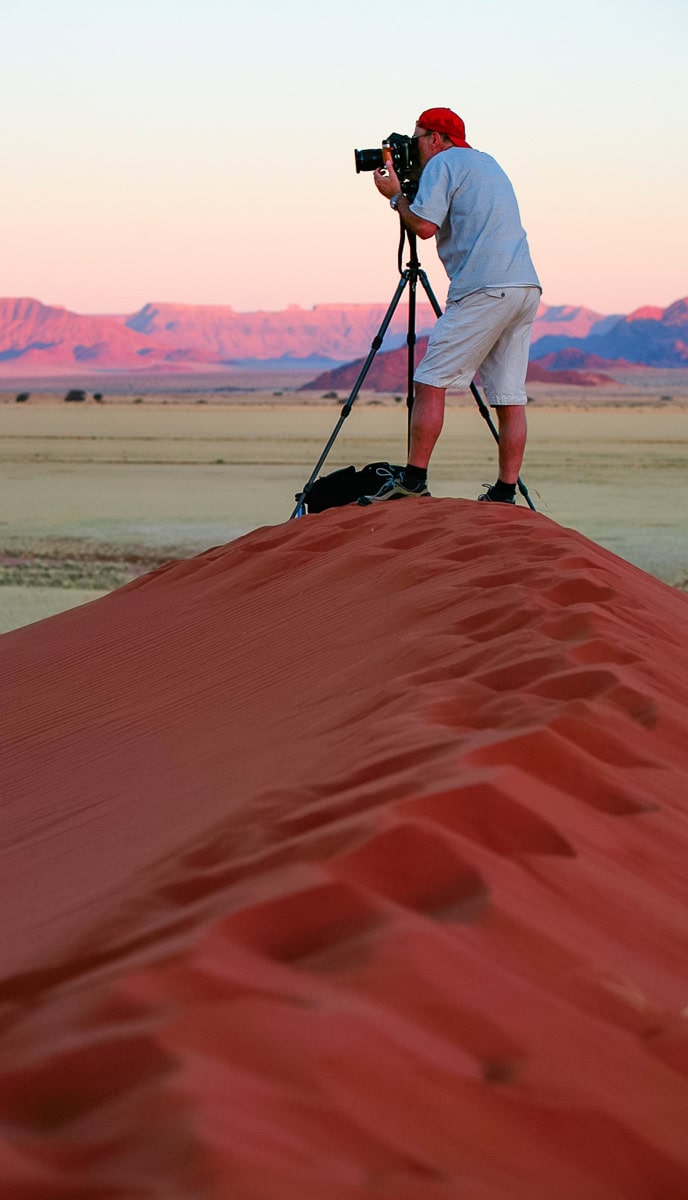 A person in white clothes and a red cap stands with a camera on a tripod, capturing a stunning photo from a red sand dune at sunrise or sunset. The majestic mountains are visible in the background, creating an ideal scene for any travel blog.