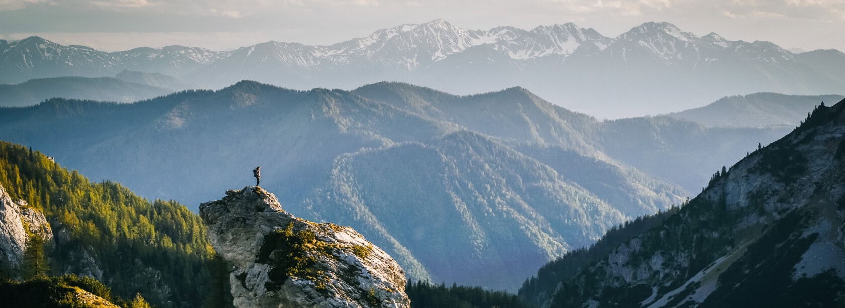 A person stands on a rocky cliff, looking out at a range of forested mountains with snow-capped peaks in the background under a partly cloudy sky, contemplating the serene beauty ideal for an inspiring travel blog.