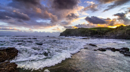 A coastal landscape at sunset features waves crashing against rocky shorelines, a distant hill reminiscent of Middle Earth, and a dramatic sky filled with clouds and sunlight.