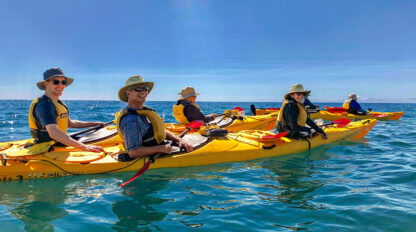 A group of people wearing hats and life jackets paddle yellow kayaks on calm, blue water under a clear New Zealand sky, with majestic mountains in the distance.