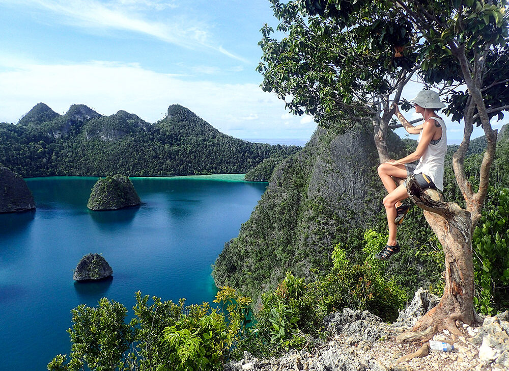 A person wearing a hat and shorts sits on a tree branch overlooking a scenic view of lush green islands surrounded by blue water under a clear sky, embracing the world's last wild frontier.