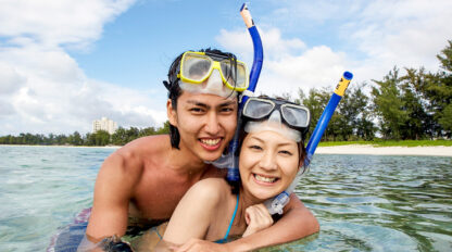 A couple wearing snorkeling gear smiles while standing in the shallow ocean with a sandy beach and greenery in the background, effortlessly embodying the joy often highlighted in snorkeling frequently asked questions.