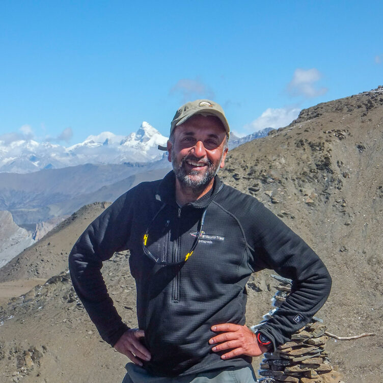 A bearded man in hiking gear and a cap stands on a rocky mountain trail, with snow-capped peaks of Kyrgyzstan in the background. This scene might as well be an invitation to explore one of the world's most breathtaking landscapes.