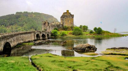 A stone castle on a small island is connected to the mainland by an arched stone bridge, with greenery and water in the foreground and wooded hills reminiscent of the Scottish Highlands in the background.