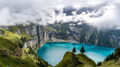 A scenic travel view of a turquoise lake surrounded by steep cliffs and green hills in Switzerland, with clouds partially covering the sky. Two trees stand prominently in the foreground on a grassy hill, capturing the essence of September's serene beauty.