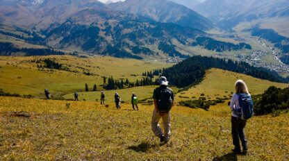 A group of hikers with backpacks descends a grassy hillside, overlooking a valley with forested areas and distant mountains under a partly cloudy sky—an invitation to explore Kyrgyzstan's breathtaking landscapes.